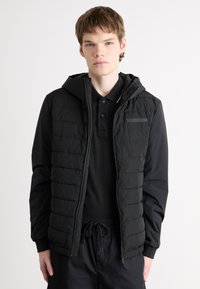 Giubbotto a vento nero con cappuccio, texture trapuntata, zipper frontale e una piccola tasca sul petto, indossato sopra una polo nera e pantaloni scuri.