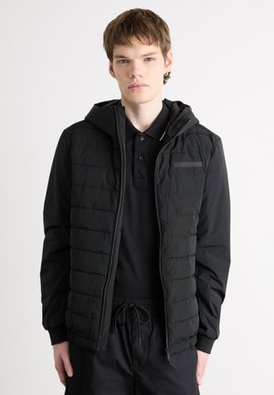 Giubbotto a vento nero con cappuccio, texture trapuntata, zipper frontale e una piccola tasca sul petto, indossato sopra una polo nera e pantaloni scuri.