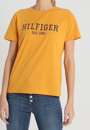 Persoon die een geel T-shirt met korte mouwen draagt met de tekst "HILFIGER EST. 1985" en een blauwe spijkerbroek met drie zichtbare voorste knopen.