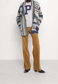 Flerfärgad stickad cardigan med geometriska mönster, grå bomullströja med marinblå "P", beige manchesterbyxor med vida ben, och svarta ankelskor.