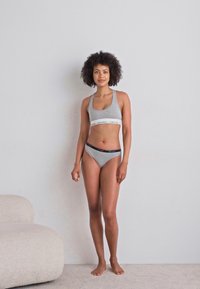 Grå sport-bh og matchende bikiniset med hvit underdel og teksturert stoff. Modellen står barføtt mot en nøytral vegg.