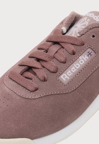 Gros plan d'une sneaker Reebok en daim mauve avec des lacets assortis et une semelle blanche, avec le logo Reebok et un petit drapeau Union Jack sur le côté.