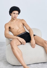 Μαύρα αντρικά μποξέρ με λογότυπο στη μέση, φορεμένα από ένα άτομο που κάθεται σε μια ανοιχτόχρωμη υφασμάτινη καρέκλα μπροστά σε ένα απαλό φόντο.