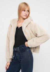 Μπεζ, αφράτο μπουφάν με φερμουάρ και κουκούλα. Συνδυάζεται με μαύρο ριμπ μπλουζάκι και τζιν παντελόνι ψηλόμεσο με ζώνη στολισμένη με σχέδιο λουλουδιού.