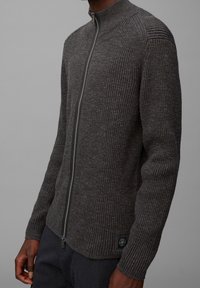 Szary sweter prążkowany z wysokim kołnierzem i pełnym zamkiem, z teksturowanym dzianiną i małą łatką z logo w lewym dolnym rogu.