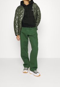 Zelená prošívaná bunda přes černou mikinu, v kombinaci se zelenými kalhotami a robustními bílými teniskami s černými akcenty.