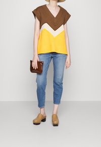 Pletený top s hnědým, krémovým a žlutým chevronovým vzorem, v kombinaci se světle modrými džíny a hnědými dřeváky, držící hnědou kabelku.