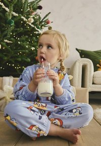 Pijamale din fleece albastre cu personaje de desene animate, ținând un pahar cu lapte cu un pai cu dungi roșii, așezate pe un podea din lemn lângă un brad de Crăciun decorat.