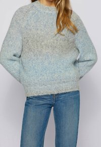 Lichtblauwe gebreide trui met een gestructureerde, vossige afwerking; heeft een ronde halslijn en raglanmouwen; gecombineerd met een blauwe spijkerbroek.