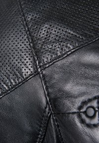 Zwarte lederen textuur met perforaties, zichtbare stiksels en een cirkelvormig hardware detail. Het materiaal toont zowel gladde als gestructureerde afwerkingen.