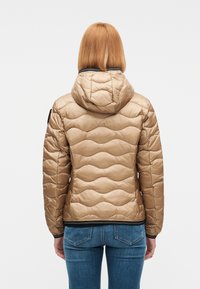 Γυναίκα με ίσια κόκκινα μαλλιά που φοράει μπεζ μπουφάν με κουκούλα και μπλε τζιν, φαινομένη από την πίσω όψη μπροστά σε απλό φόντο.