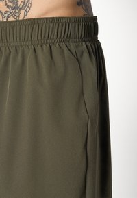 Close-up van olijfgroene elastische tailleband broeken met zijzak en gedeeltelijk zichtbare getatoeëerde torso op een witte achtergrond.