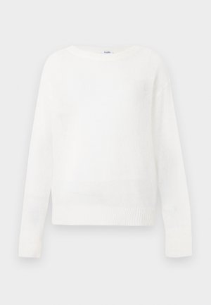 Femme portant un pull blanc, un pantalon large foncé, des ballerines noires, tenant un grand sac en cuir noir sur fond blanc uni.