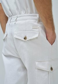 Pantalones cargo blancos con una textura suave, dos bolsillos traseros con solapas y botones, y una pequeña etiqueta de la marca en la cintura.