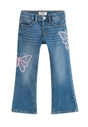 Ung pige med krøllet hår iført en hvid t-shirt, blå jeans med pink sommerfuglemotiver og hvide sneakers, står med hænderne i lommerne.