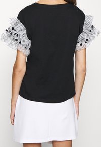 Černé tričko s průhlednými rukávy s puntíky. Rukávy mají bílé volány s černými tečkami, což přidává texturu a vizuální zajímavost.