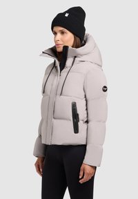 Lys grå pufferjakke med en høj krave, sorte lynlåse og en sort logo-patch. Båret med en sort ribbet hue.