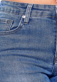 Vaqueros de mezclilla azul de talle alto, con un bolsillo frontal y detalle de botón metálico. La tela tiene una textura clásica y un ligero desgastado.