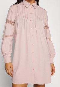 Helles rosa Hemdkleid mit einem kragenlosen Ausschnitt, vorderen Knöpfen, gefälteltem Oberteil und Spitzenakzenten an den Ärmeln. Lange, locker sitzende Ärmel.