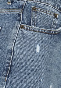 Tela de denim de color azul claro con zonas desgastadas, costuras visibles y detalles de herrajes metálicos. Incluye un pequeño bolsillo trasero y acentos de botones.