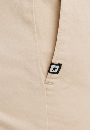 Beežist kangast tasku nähtava õmblusega ja väikese musta-valge ruudukujulise sildiga, millel on ääre lähedal must täht.