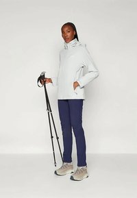 Lys grå vanntett jakke med høy krage, kombinert med marineblå bukser og beige turstøvler. Holder en svart trekkingstokk.