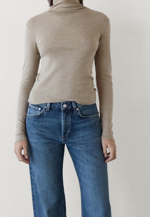 Personne portant un pull beige à col roulé à manches longues et un jean bleu taille haute sur un fond clair uni.