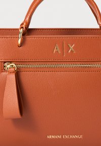 Sac à main en cuir texturé orange avec quincaillerie dorée, doté d'une fermeture éclair, des initiales du logo "A|X" sur le devant, et d'un onglet décoratif suspendu.