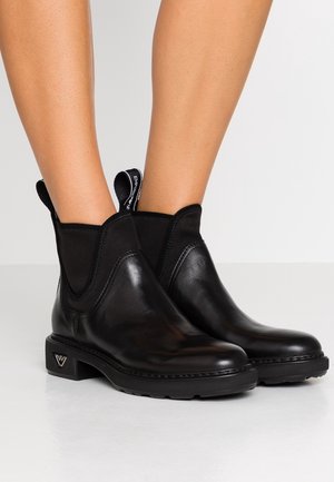 Botas tobilleras de cuero negro con paneles laterales elásticos y tiradores con marca, usadas sobre piernas desnudas contra un fondo claro y liso.