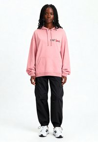 Felpa rosa con cappuccio, lacci e tasca frontale, con la scritta "SWEET" ricamata in rosso. Abbinata a pantaloni cargo neri e sneaker bianche.