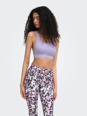 Jeune femme aux longs cheveux bouclés, portant un soutien-gorge de sport lavande et des leggings colorés à motifs, debout devant un fond uni.