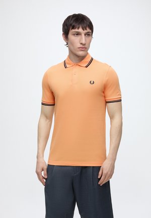 Mężczyzna ubrany w pomarańczową koszulkę polo z krótkim rękawem, z czarną lamówką na kołnierzu i rękawach, w połączeniu z ciemnoszarymi spodniami, stojący na białym tle.