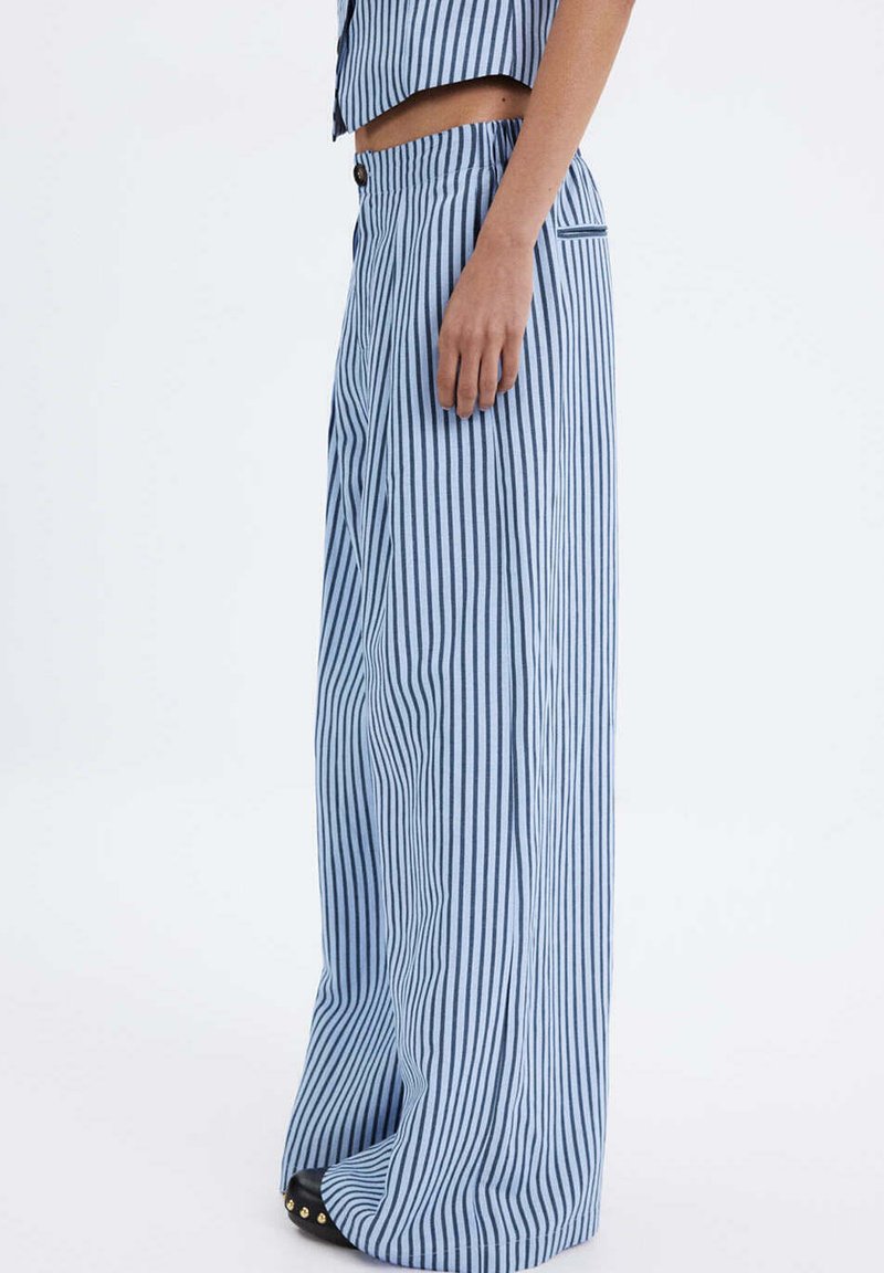 Femme portant un pantalon taille haute à jambes larges avec des rayures verticales bleues et blanches, un crop top assorti et des chaussures noires avec des clous dorés.