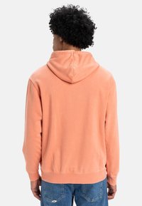 Sudadera de melocotón con capucha ajustable, textura suave, puños y cinturilla de canalé, combinada con jeans de mezclilla azules. La vista trasera muestra los detalles de las costuras.
