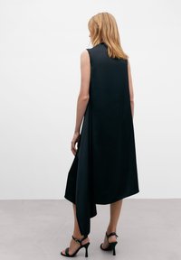 Robe sans manches en satin noir avec une hem asymétrique, texture lisse, col haut, et associée à des talons strappés noirs.