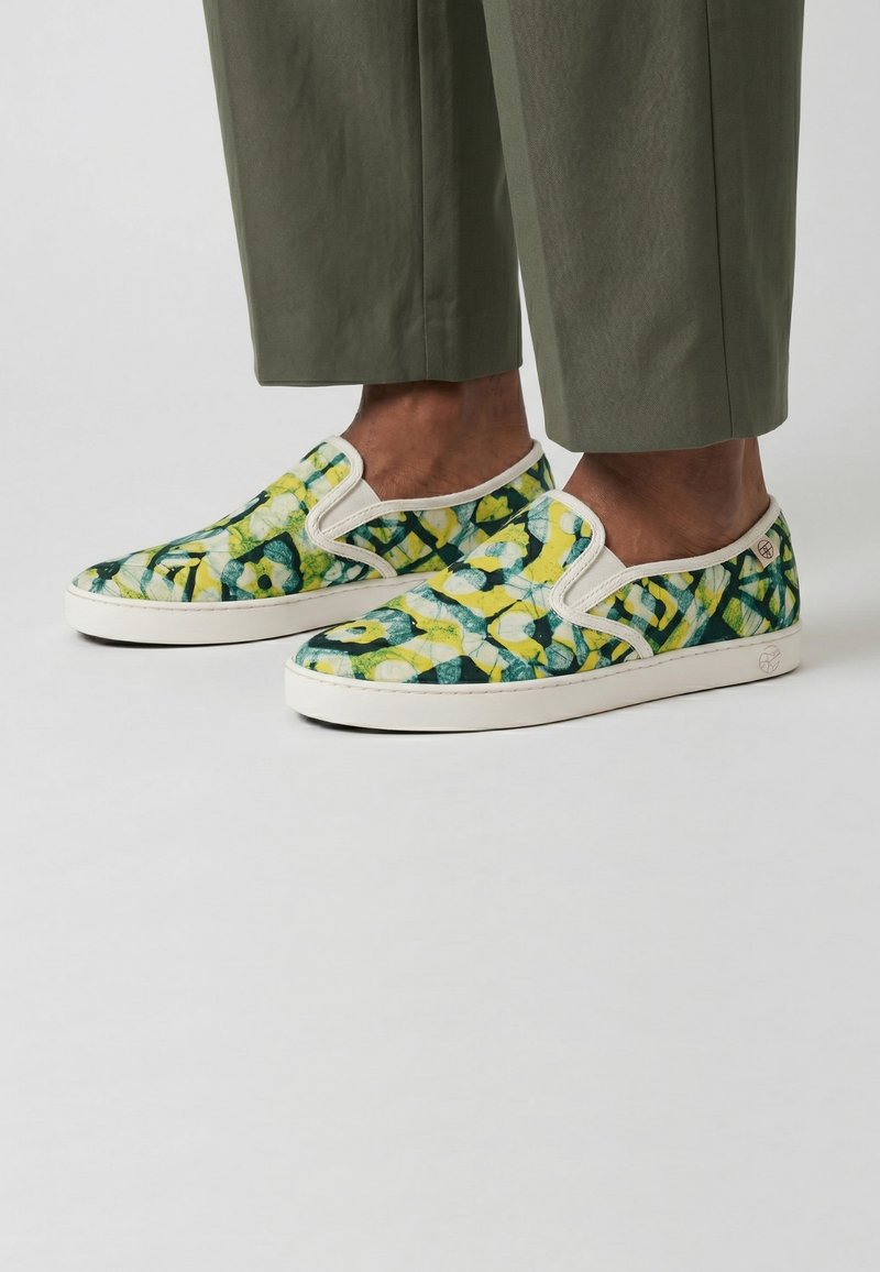 Pieds chaussés de slip-on aux motifs floraux abstraits verts, jaunes et blancs, associés à un pantalon court vert olive sur un fond uni.