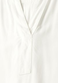 Blouse blanche avec un décolleté en V, présentant une patte de boutonnage cousue. Fabriquée en tissu léger avec une texture lisse et des détails minimaux.