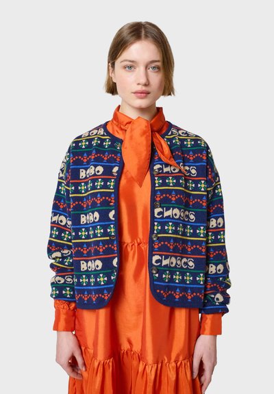 Cárdigan de punto azul con rayas estampadas en rojo, verde y blanco, con cuello redondo y cierre de botones, llevado sobre un vestido naranja brillante.