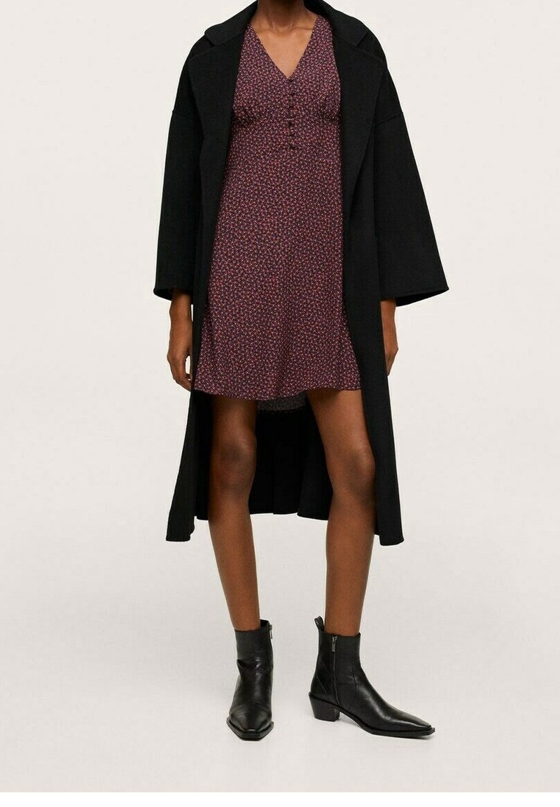 Femme portant un long manteau noir sur une courte robe bordeaux à motifs et des bottines noires sur un fond clair uni.