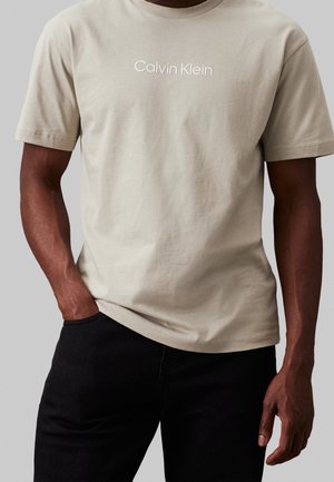 Hombre con camiseta beige de manga corta Calvin Klein y pantalones negros, con una mano en el bolsillo frente a un fondo gris liso.