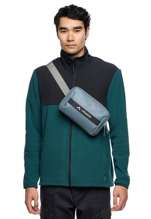 Graue Umhängetasche mit schwarzen Akzenten, glatter Textur, rechteckiger Form und verstellbarem Riemen, getragen über einer grünen Fleecejacke.
