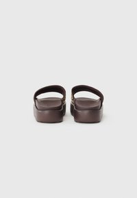 Brown slip-on sandals with a textured bottom, wide back strap, and smooth upper. Features a small decorative buckle on each side.