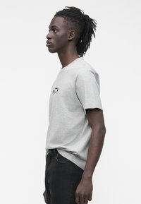 Pilkas medvilninis marškinėlis su apvalia iškirpte, trumpomis rankovėmis ir mažu raudonu logotipu ant krūtinės. Dėvimas su juodais džinsais, matomas iš šono.