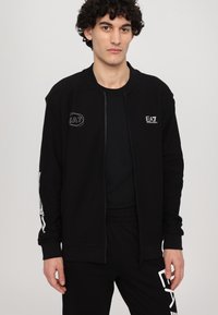 Black zip-up jacket with textured fabric, featuring white EA7 logo accents on the chest and sleeve. Paired with a black t-shirt and pants.
