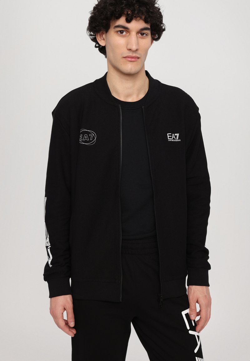 Chaqueta negra con cremallera de tela texturizada, con detalles del logo blanco de EA7 en el pecho y la manga. Combinada con una camiseta y pantalones negros.
