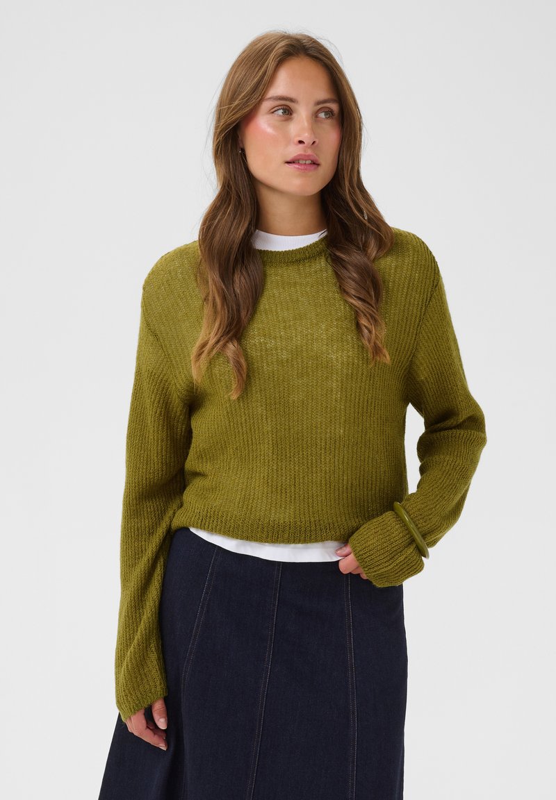 Femme portant un pull en tricot vert olive sur un haut blanc et une jupe bleu foncé, debout devant un fond clair uni.