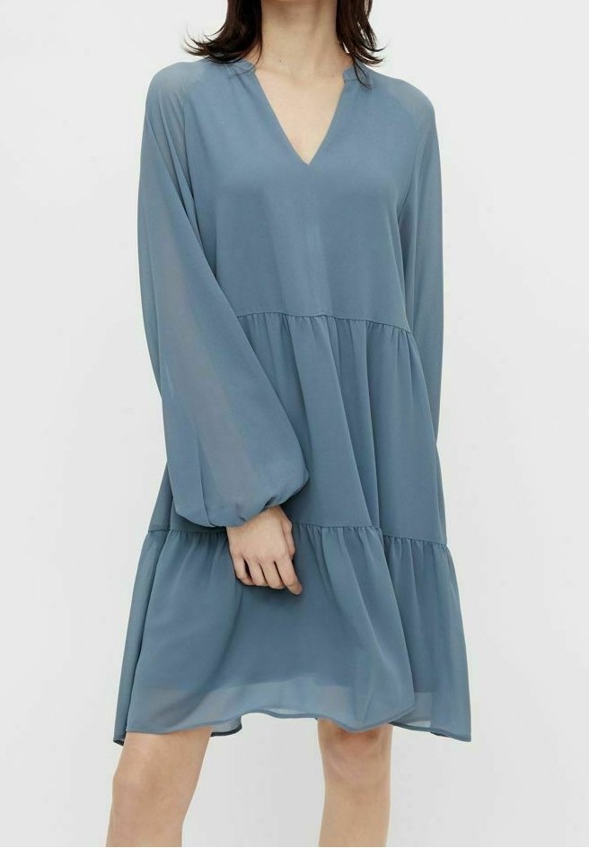 Femme portant une robe bleue ample à manches longues, longueur genoux, avec un col en V et une jupe à volants, debout devant un fond uni.