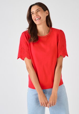 Donna sorridente con capelli castani che indossa una camicetta rossa brillante con maniche smerlate ricamate e jeans azzurri chiari, su uno sfondo uniforme.