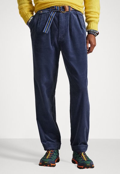 Marineblaue Cordhosen mit hoher Taille, Falten und einem bunten gestreiften Gürtel. Kombiniert mit strukturierten, mehrfarbigen Sportschuhen.