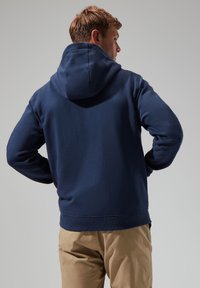Navy blue hoodie with a large back hood, fleece texture, and ribbed cuffs. Paired with light beige pants.